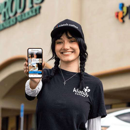 Barista holding a smartphone displaying the Klatch Coffee app outside of a Sprouts Farmers Market and Klatch location.
