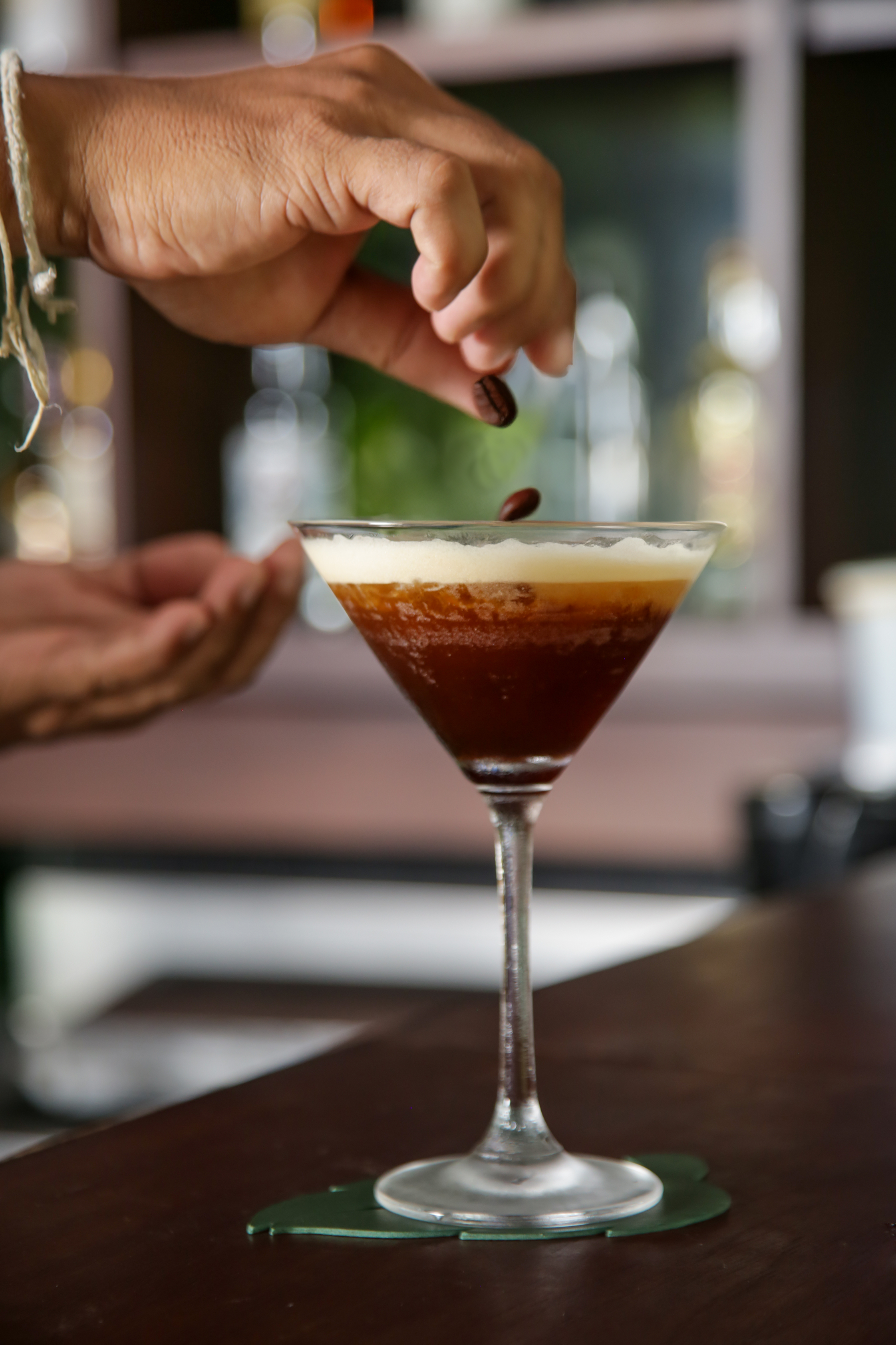 Cocktail glass with a dark liquid being garnished with a coffee bean on a wooden surface.