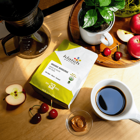 Organic Honduras Mano De Mujer Fair Trade coffee bag on wooden table, glass ethoz pour-over with white mug with coffee. Fruit of flavor notes of Coffee plant and  red apples, one cut in wedge with two cherries on tray with half apple and cherries laying on table at the bottom of the bag next to glass bowl of caramel and mug of coffee.