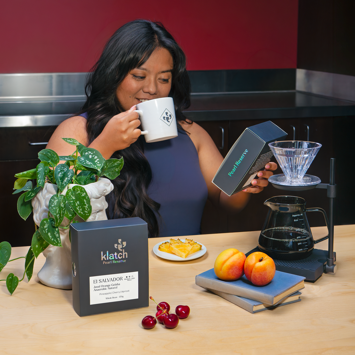 Person enjoying a cup of coffee while holding a Klatch Coffee Pearl Reserve El Salvador Jasal Orange Geisha box. A pour over brewer, carafe, fresh pineapple, apricots, and cherries are arranged on the table to highlight the coffee's tasting notes.