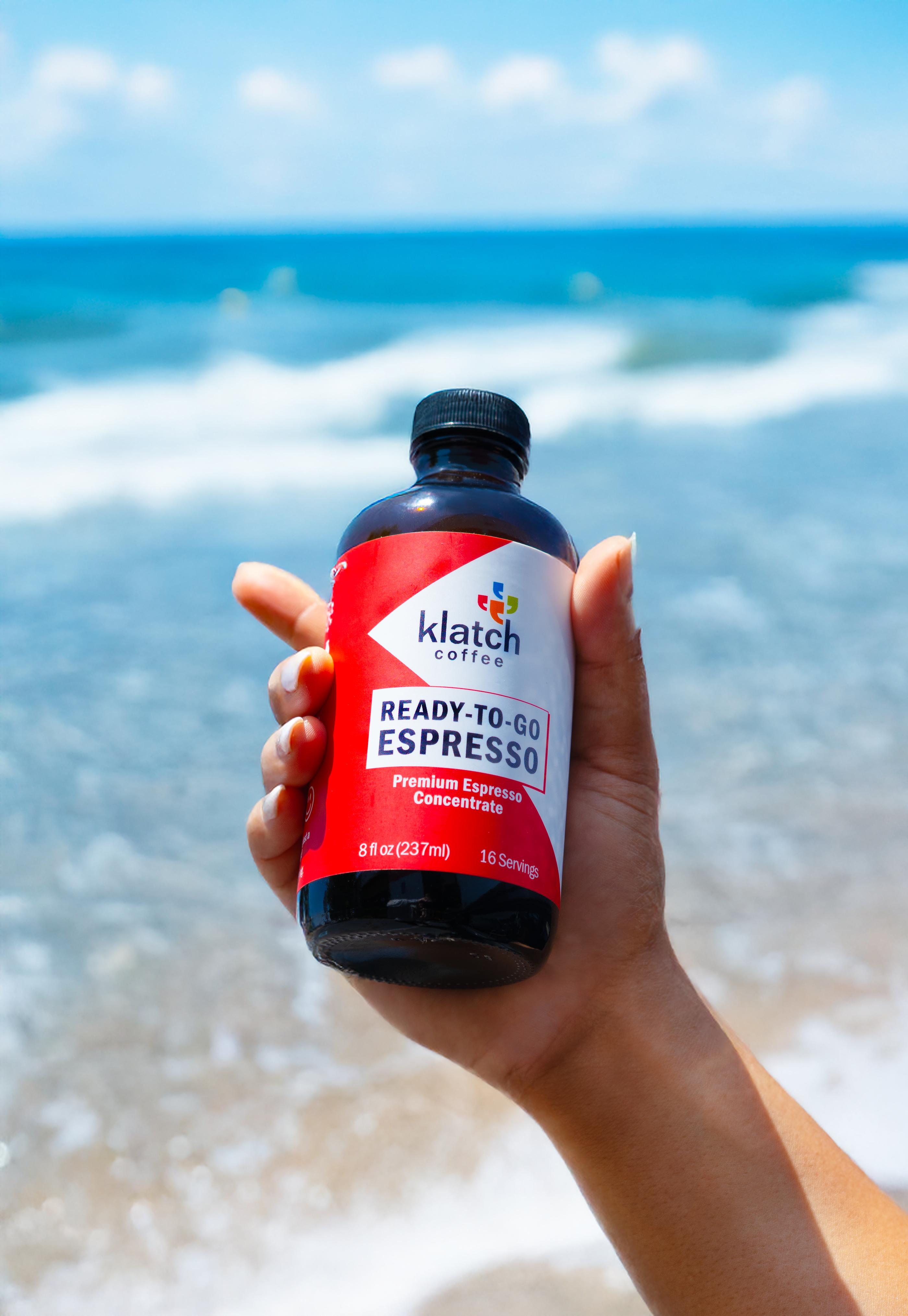 Hand holding an 8oz bottle of ready to go espresso in front of a beach. 