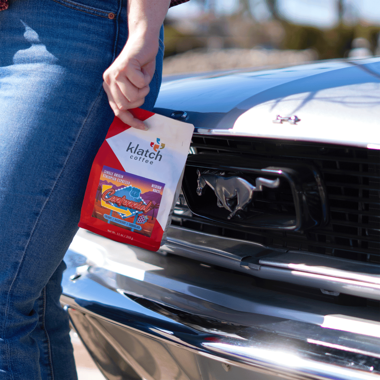 Person holding a Klatch Coffee Centennial bag in front of a Ford Mustang.