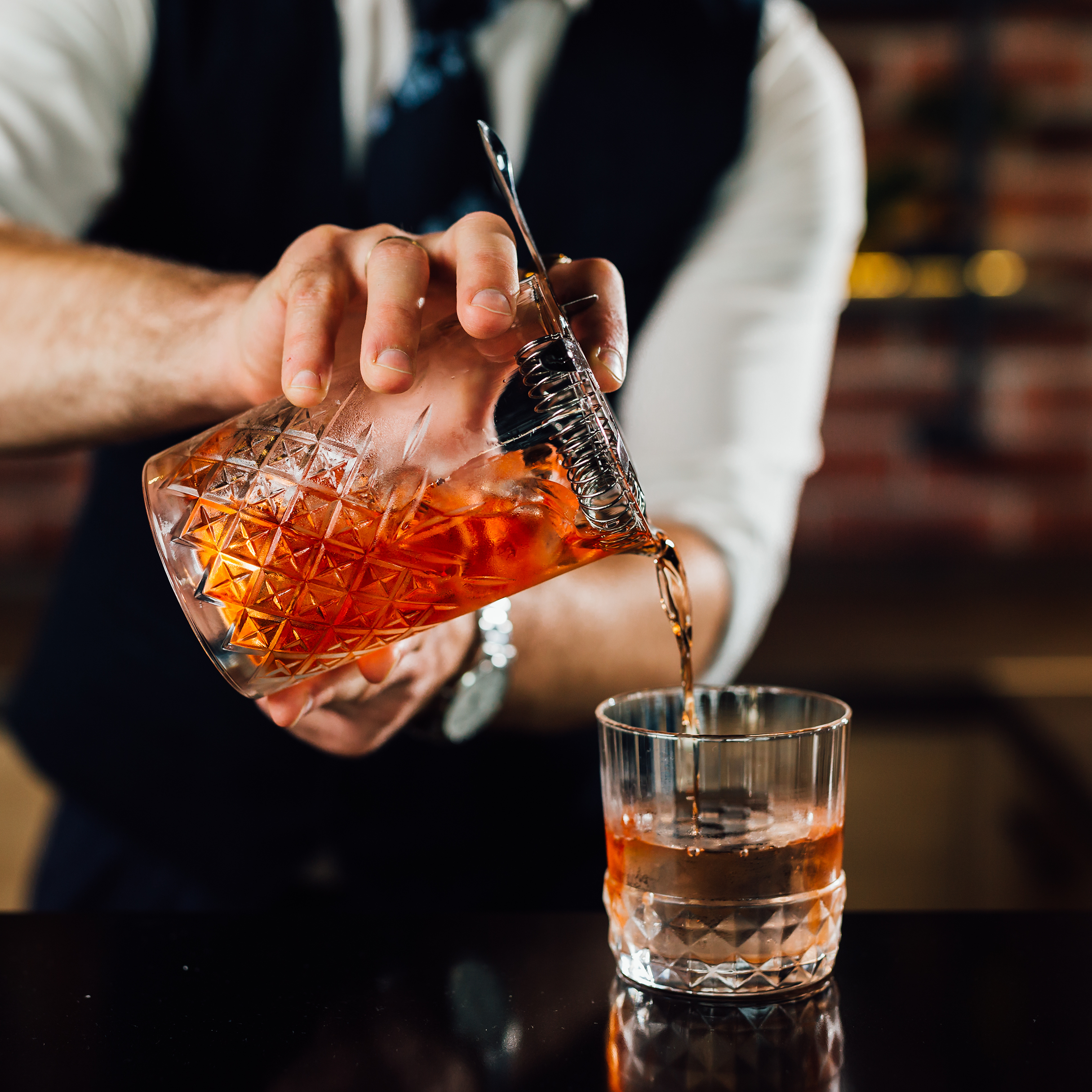 Bartender pouring a cocktail from a glass shaker into a glass.